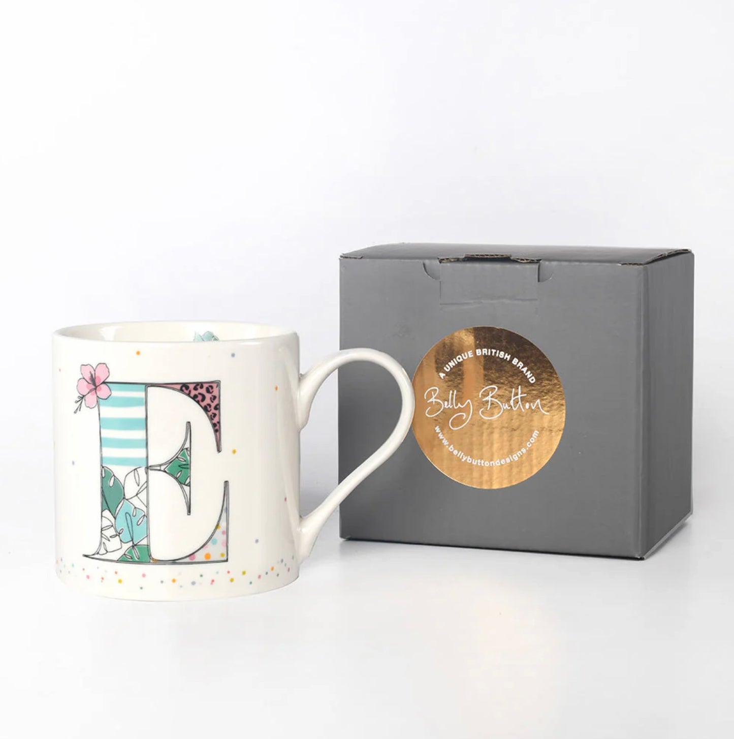 A white ceramic mug with a multicolor floral and animal pattern and a large gold letter 'E' in the center. The mug is displayed next to a gray presentation box with a foil stamp on the lid.