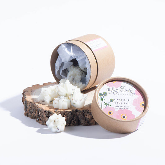 A collection of flower-shaped scented soy wax melts spilling out of an open cylindrical container with a label that reads 'Belly Button Designs - Pink Cosmos'. The wax melts are placed on a wooden slice.