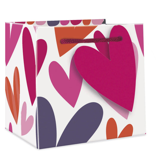 A gift bag with a woven paper handle and a design of large hearts in shades of pink, purple, and orange on a white background. The bag has a closure with a red ribbon.