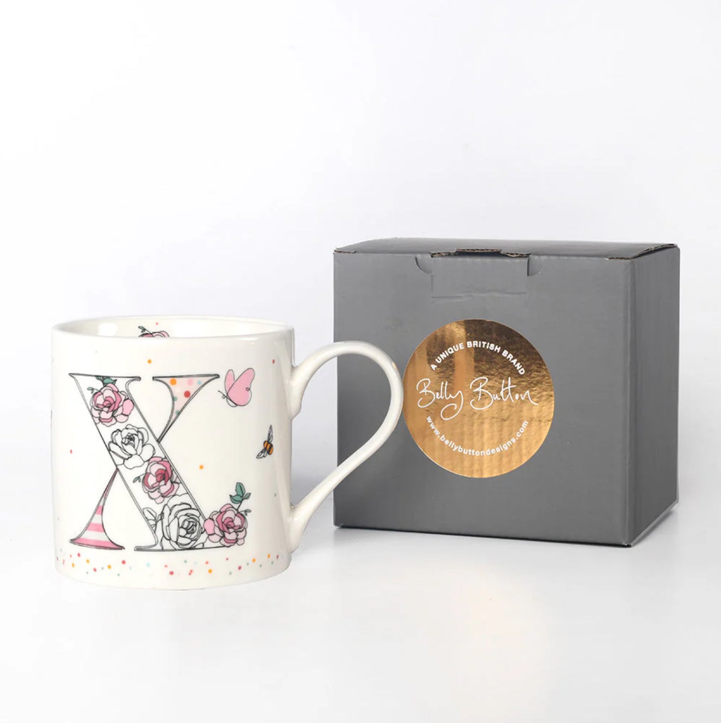 A white ceramic mug with a floral and monogram initial 'X' design, displayed in front of a grey presentation box with a foil stamp on top.