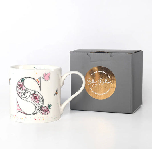 A white ceramic mug with a floral and whimsical design, featuring an ornate letter 'S' in the center, presented next to a gray presentation box with a logo on it.