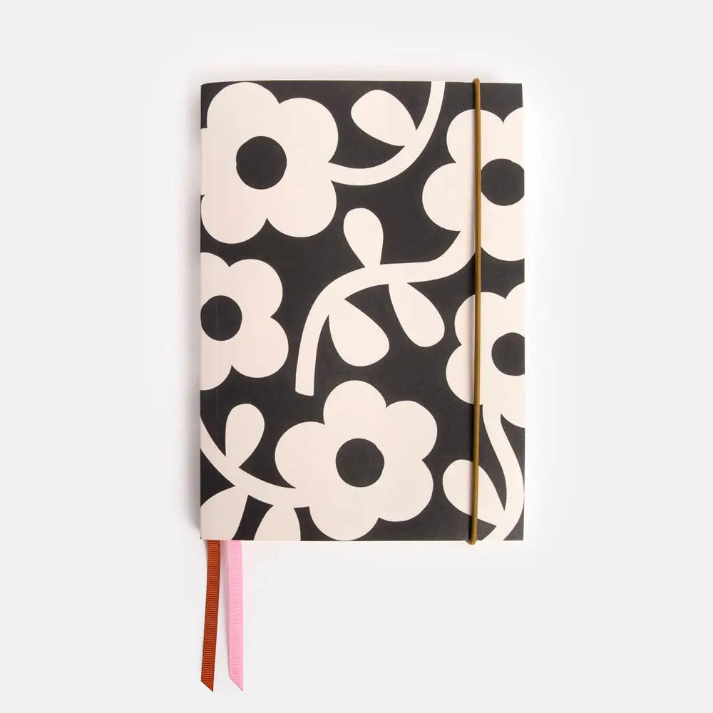 Caroline Gardener A5 notebook with black cover featuring bold white floral pattern, pink and burnt orange ribbon bookmarks, and gold elastic closure.