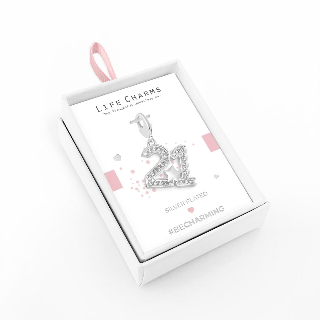 A silver-plated number 21 charm with clear zirconia stones presented in a white box with 'LIFE CHARMS' branding, attached to a pink ribbon.
