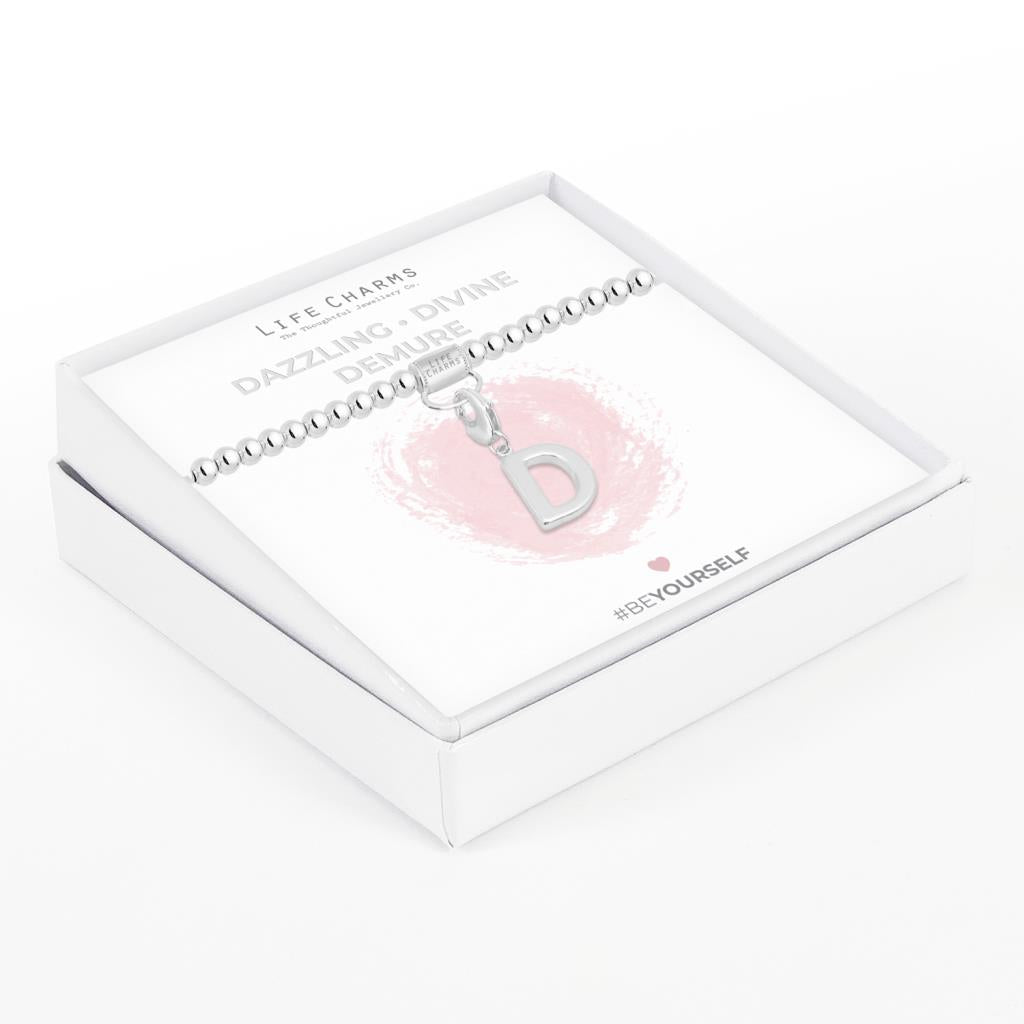 Initial charm bracelet with the letter D, presented on a silver foiled card with the text 'DAZZLING • DIVINE • DEMURE' and the brand name 'LIFE CHARMS'. The bracelet is silver, and the card has a pink background with a heart shape.