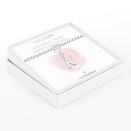 Initial charm bracelet with the letter K, presented on a silver foiled card with the text 'KISSABLE • KIND • KNOWLEDGEABLE' and the brand name 'LIFE CHARMS'. The bracelet is silver, and the card has a pink background with a heart shape.
