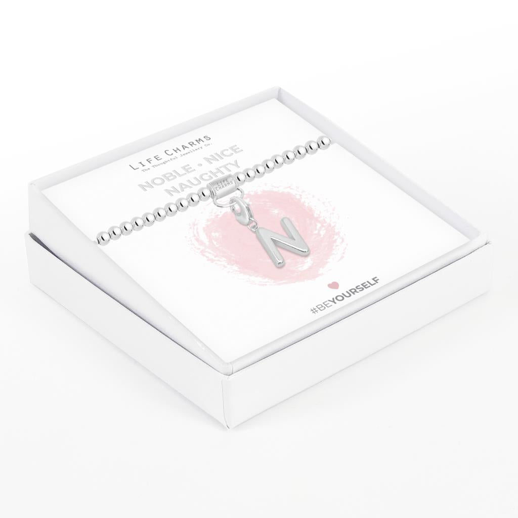 Initial charm bracelet with the letter N, presented on a silver foiled card with the text 'NOBLE • NICE • NAUGHTY' and the brand name 'LIFE CHARMS'. The bracelet is silver, and the card has a pink background with a heart shape.