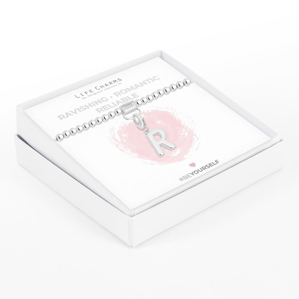 Initial charm bracelet with the letter R, presented on a silver foiled card with the text 'RAVISHING • ROMANTIC • RELIABLE' and the brand name 'LIFE CHARMS'. The bracelet is silver, and the card has a pink background with a heart shape.
